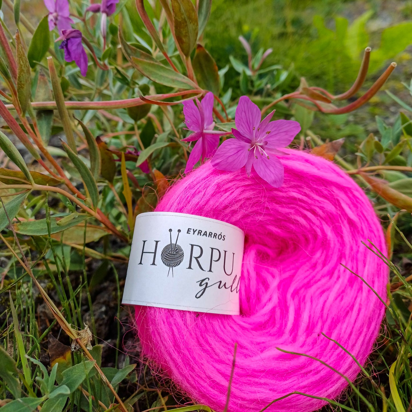 Hand-dyed Icelandic lopi yarn inspired by Arctic riverbeauty placed next to pink wildflowers in nature